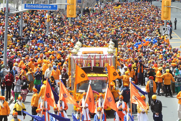 Hundreds walking in a parade, behind a bus with a temple-like structure atop and several men in front of it, holding saffron-color flags.