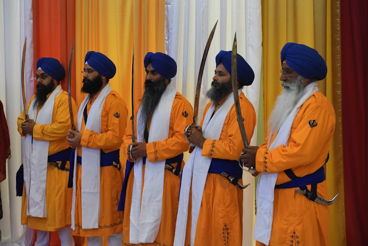 Five men with turbans and flowing beards, dressed in long orange shirts and white scarfs, holding swords in their hands.