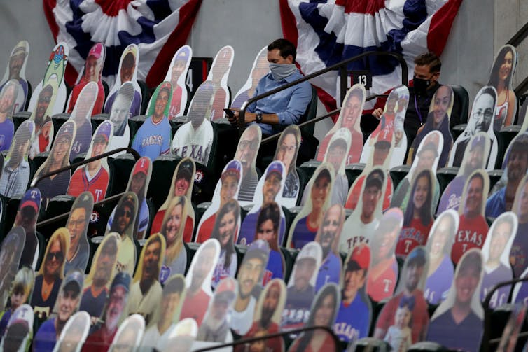 Colorful cutouts of smiling fans are placed in seats. In this sea of fake fans, two men wearing face masks take notes and look at their phones.