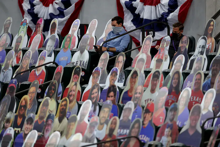 There are colorful cutouts of smiling fans on the seats. In this sea of fake fans, two men wearing face masks are taking notes and looking at their phones.