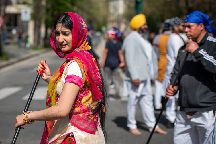A woman, dressed in a bright pink and white shirt, holds a broom as she sweeps the street, accompanied by a man in a blue turban behind her, who is also holding a broom.