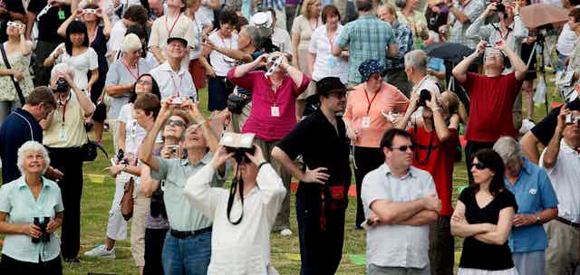 Des personnes debout regardent le ciel en portant des lunettes d'éclipse, tandis que des personnes dans la foule tiennent des appareils photo.