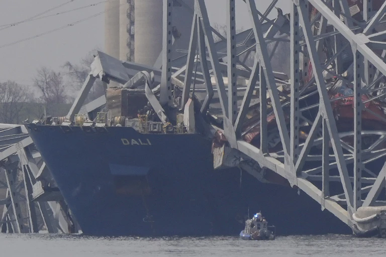 the front of a container ship floats with large chunks of the bridge it crashed into lying on top