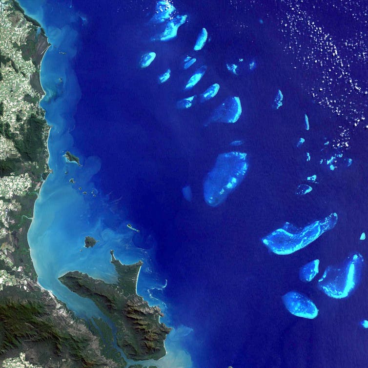 An overhead view of a deep blue ocean with brighter islets off a green coast.