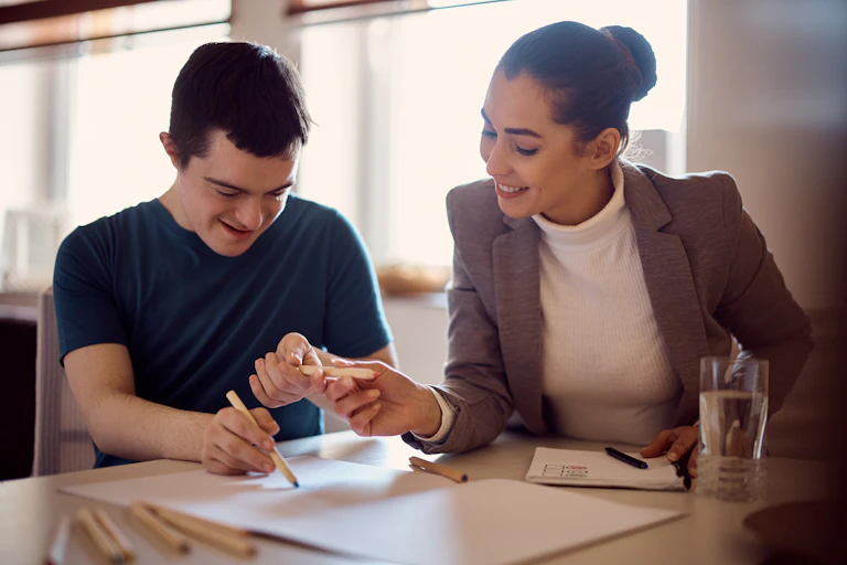 Two people sit at a desk. The woman is helping the man with drawing.