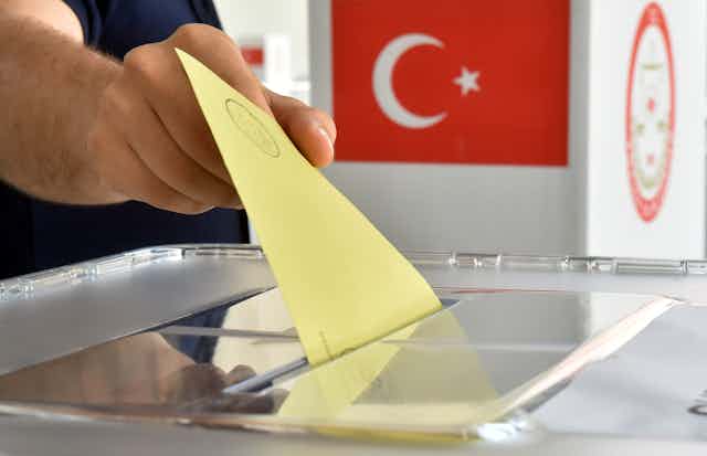 A man puts a ballot into a ballot box in front of the national flag of Turkey.