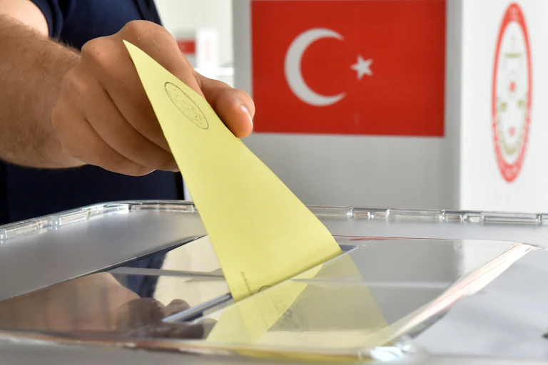 A man puts a ballot into a ballot box in front of the national flag of Turkey.