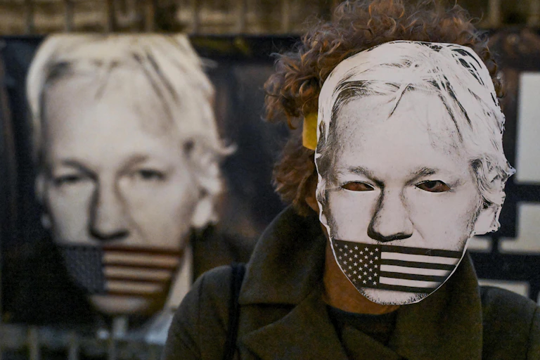 A person wearing a paper mask depicting Julian Assange's face with an american flag over his mouth