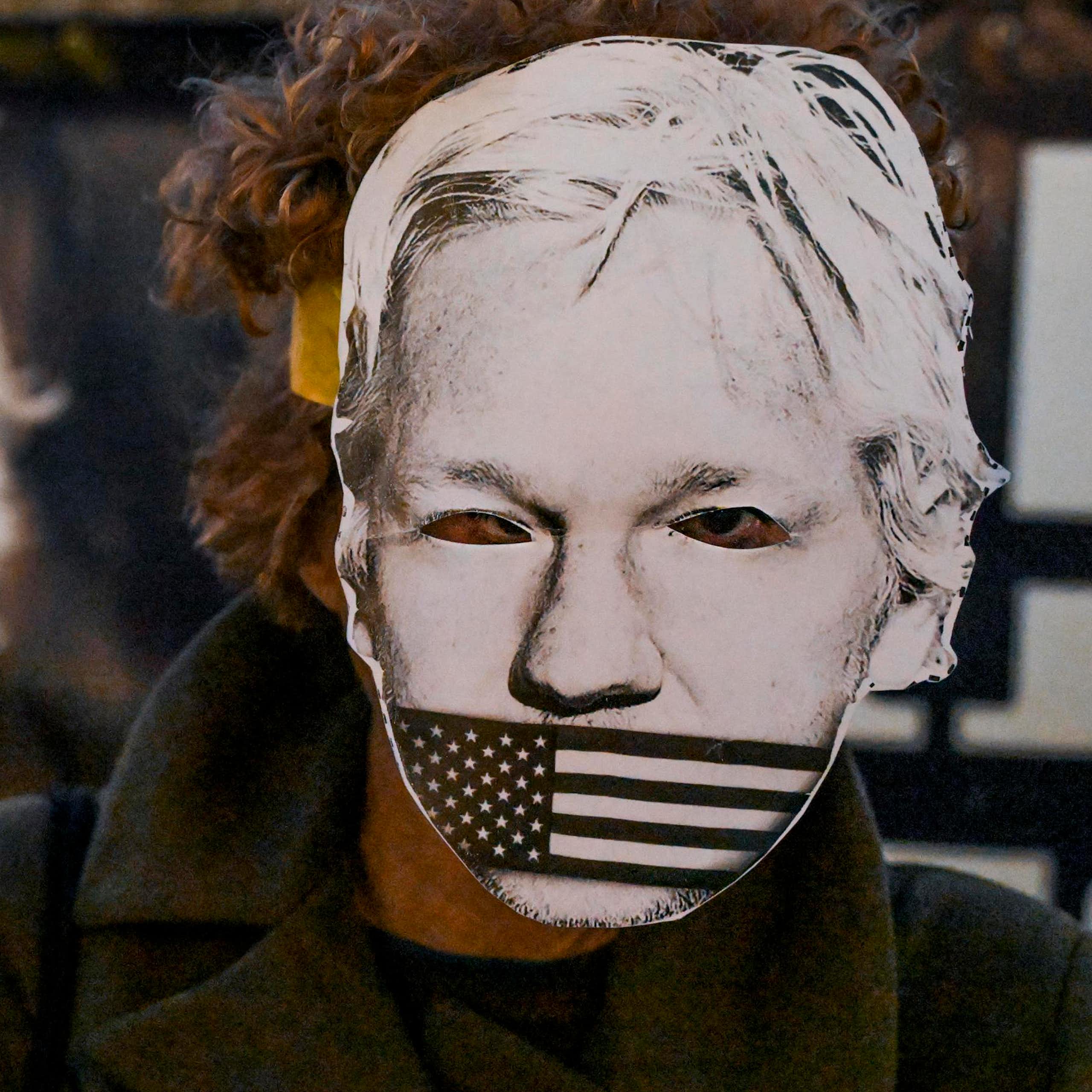 A person wearing a paper mask depicting Julian Assange's face with an american flag over his mouth