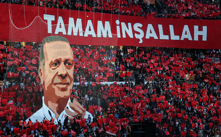 A stand full of people holding red placards and a large picture depicting Turkish president, Recep Tayyip Erdoğan.