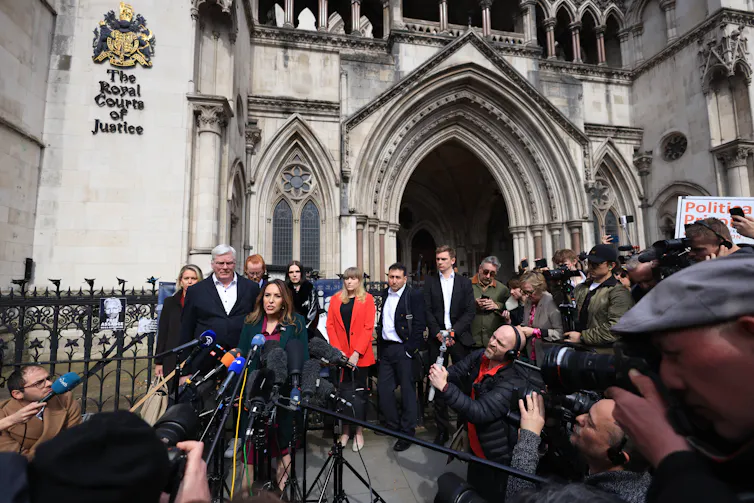 A group of people speaking into press microphones and surrounded by reporters, with the grand Royal Courts of Justice building behind them