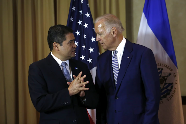Two men in suits talk in front of flags.