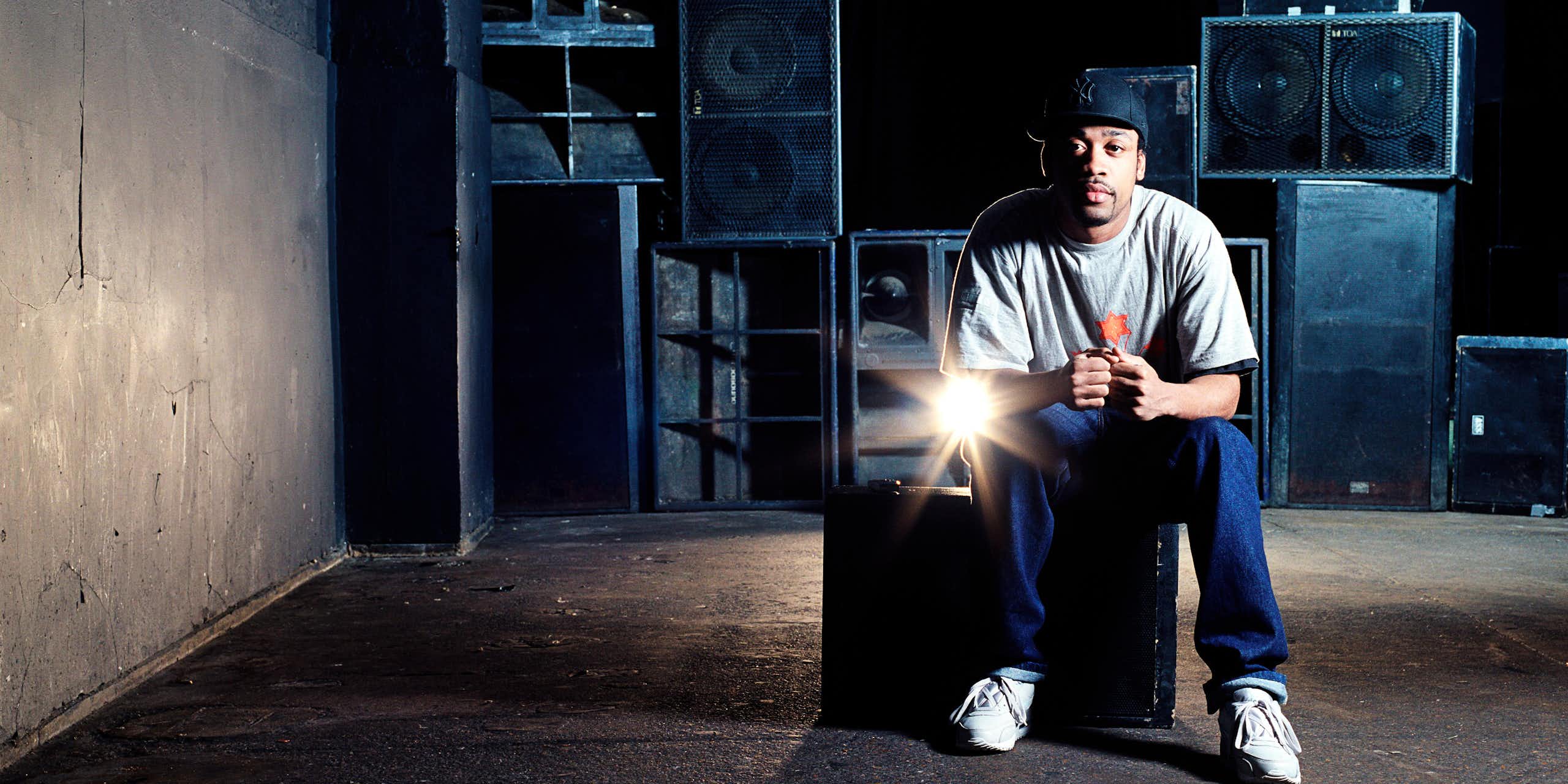 Grime artist Wiley sitting on a speaker in front of a wall of speakers. 