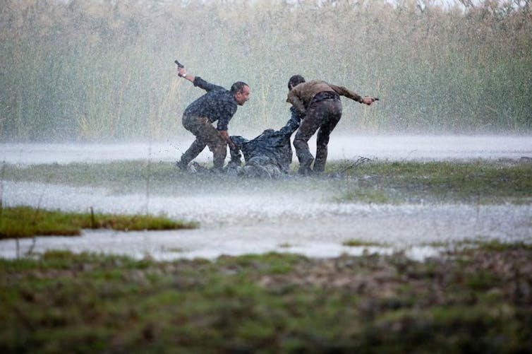 Two men drag another one through the grass on a very rainy day.
