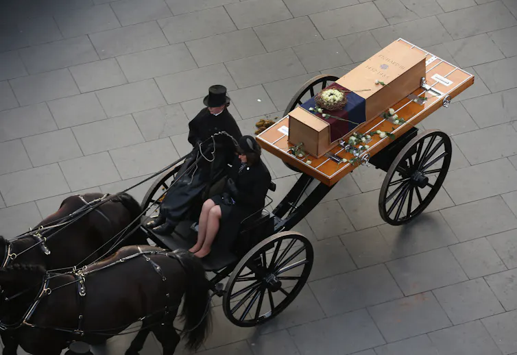 Carroça puxada por cavalos com duas pessoas vestidas de preto na frente, puxando um caixão marcado como 'Ricardo III, 1452-1485'