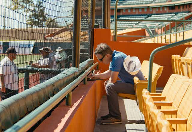 Man wearing sunglasses sitting in an empty stadium takes notes.