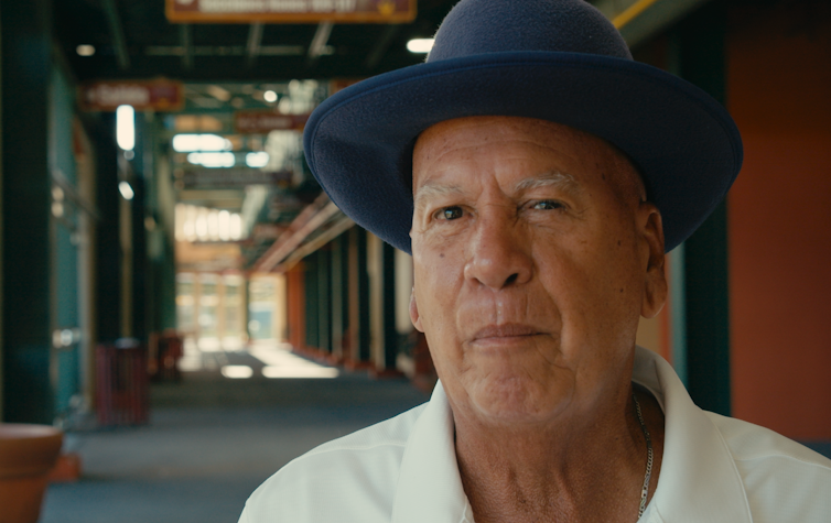Portrait of elderly man wearing blue felt hat and white polo shirt.