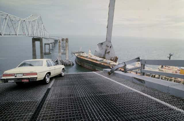 a car sits at an angle on a bridge roadway at the edge of a collapsed section with a cargo ship in the background