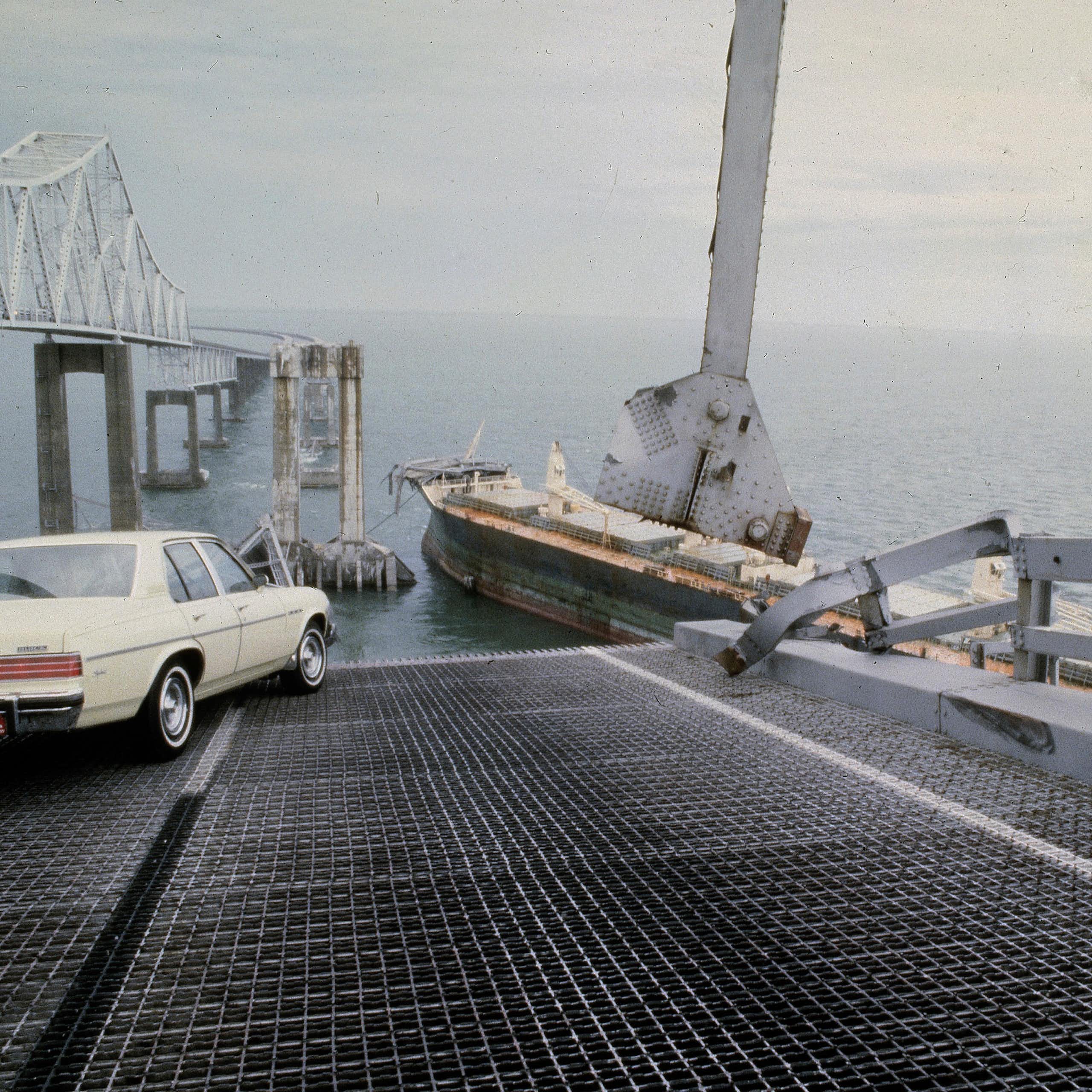 a car sits at an angle on a bridge roadway at the edge of a collapsed section with a cargo ship in the background