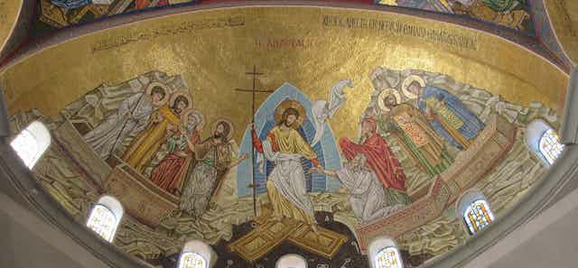 Um mosaico com fundo dourado, seis figuras com halos, Jesus segurando uma cruz no meio e duas pessoas que ele está ajudando a sair de caixões.
