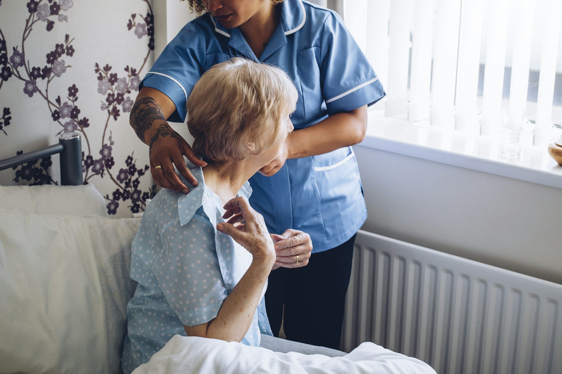 A young woman helps an elderly woman, who is seated in bed, put a shirt on