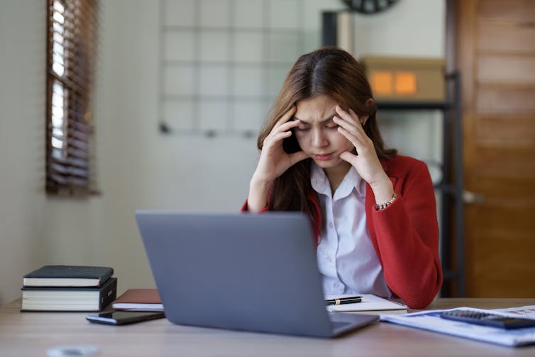 A young woman with an exhausted look on her face rests her head in her hands while sitting at a desk in front of an open laptop