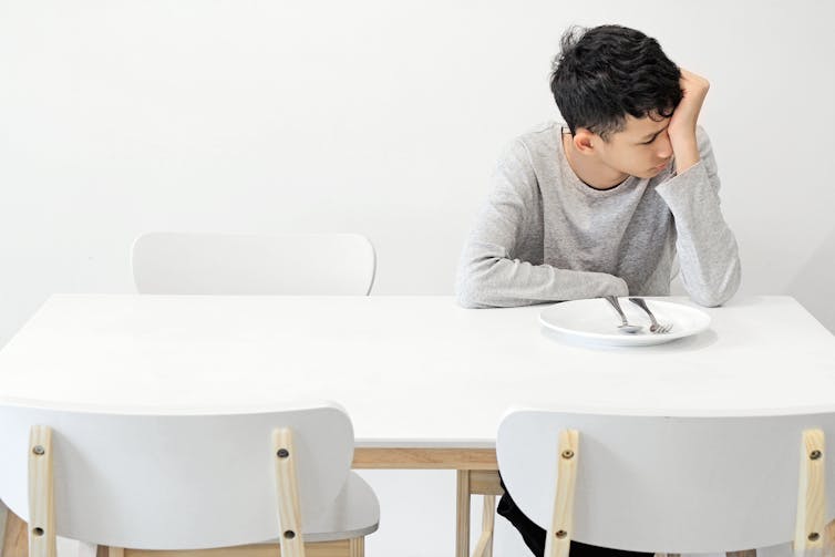 A teen boy sitting at a white table with an empty white plate on it, resting his head on his hand