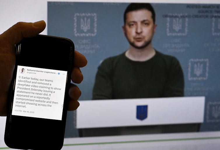 A hand holds a smartphone with text on it in front of a screen, a man stands in front of a lectern