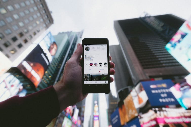 A person holding a smartphone displaying an instagram profile at a high angle against a city backdrop.