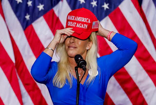 Marjorie Taylor Greene putting on a red hat with the slogan 'make America great again' written on the front, standing in front of US flags. 