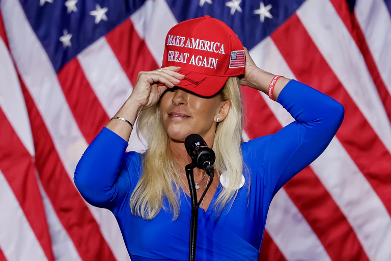 Marjorie Taylor Greene putting on a red hat with the slogan 'make America great again' written on the front, standing in front of US flags.