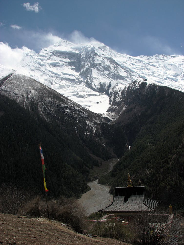 Snowcapped mountain rising into a blue sky, with a thin flagpole with prayer flags and a pagoda in the foreground