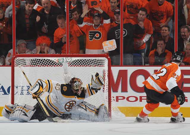 Fans dressed in orange and black watch from the stands behind protective glass as a goalie in a white, black and gold jersey strains to catch a puck with his glove.