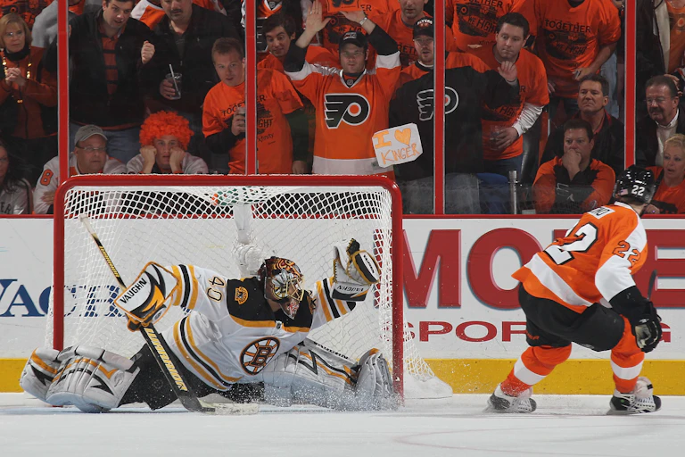 Fans dressed in orange and black watch from the stands behind protective glass as a goalie in a white, black and gold jersey strains to catch a puck with his glove.
