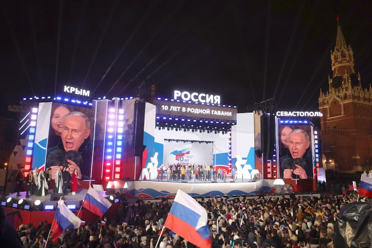 A crowd watches Vladimir Putin on video screens at a celebration of 10 years since Crimea was annexed to Russia