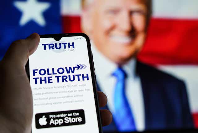 The Truth Social app open on a smartphone with a picture of Donald Trump in the background.