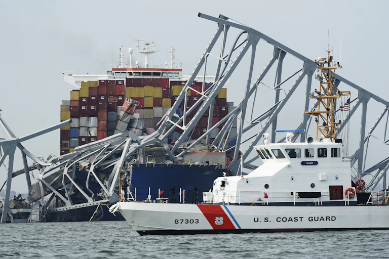 a ship tangled up in a bridge, a white US coast guard boat in front of it