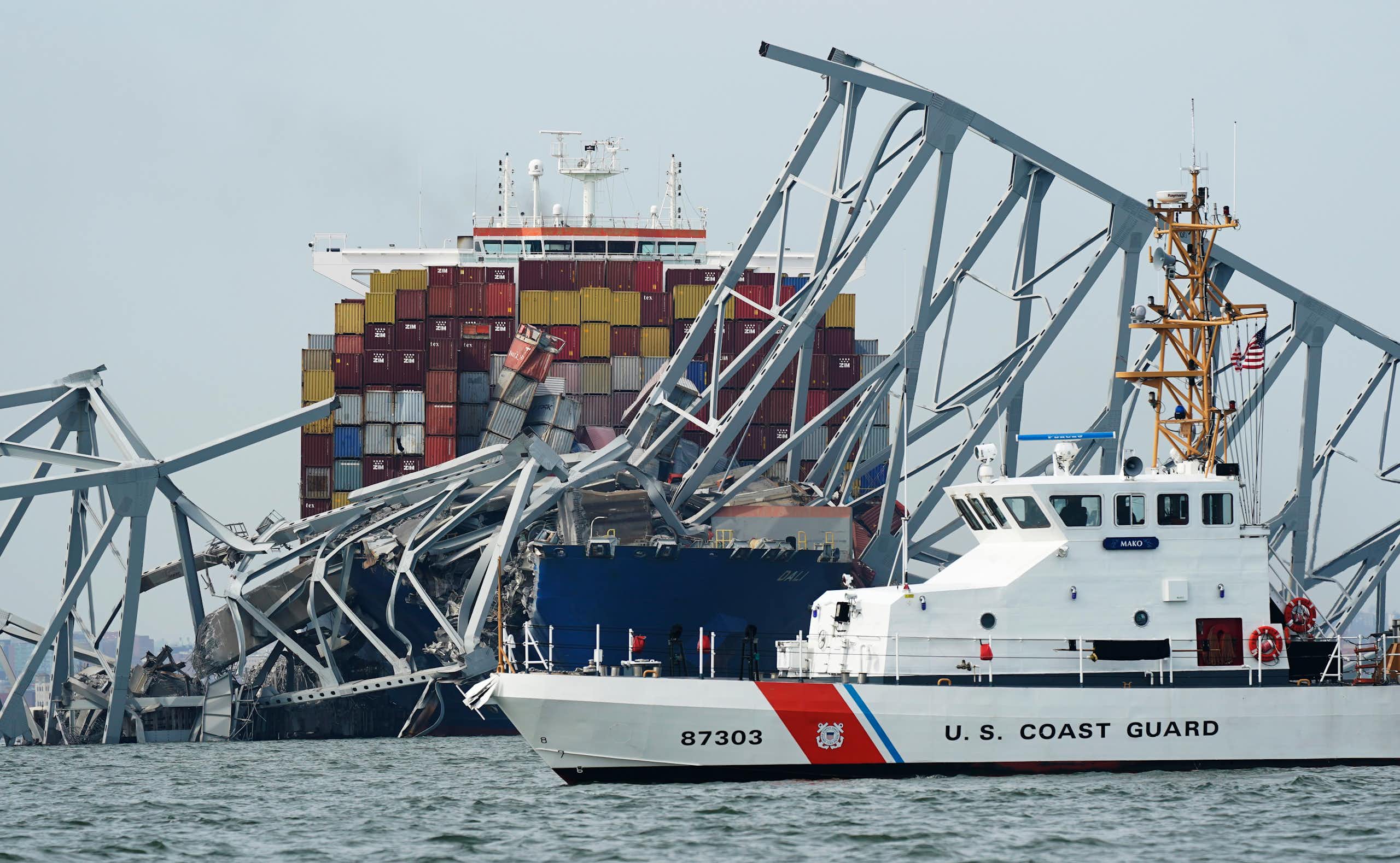 a ship tangled up in a bridge, a white US coast guard boat in front of it