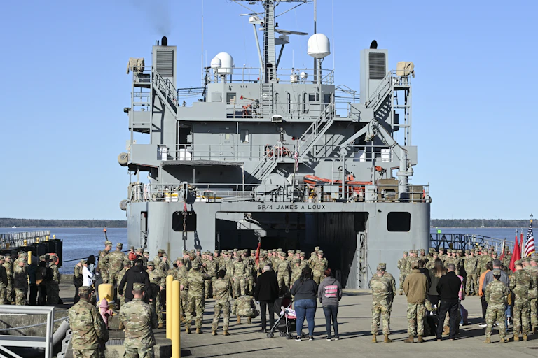 A large group of people in camouflage uniform stand on the platform of a big grey Navy boat.