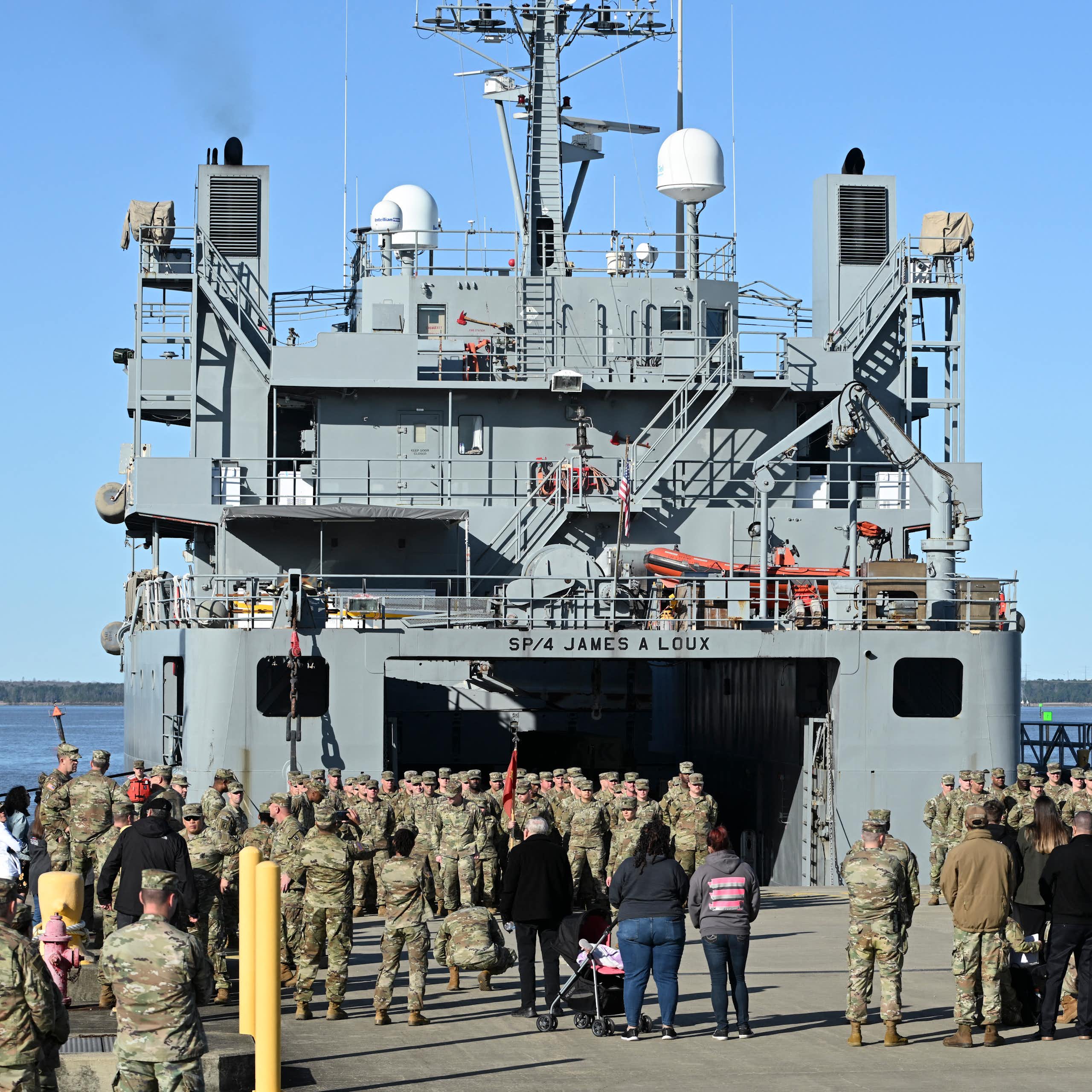 A large group of people in camouflage uniform stand on the platform of a big grey Navy boat.