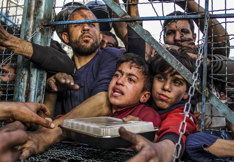 Two young boys hold a food container through a hole in a fence, surrounded by other men and hands reaching toward them.