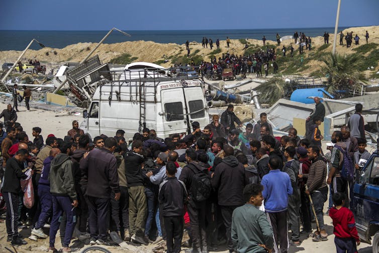 A large crowd of people huddle together near a white truck and some rubble, with the ocean in the distance.