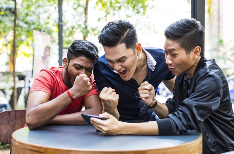 Three young men pump their fists as they look at a cell phone while seated around a round table.
