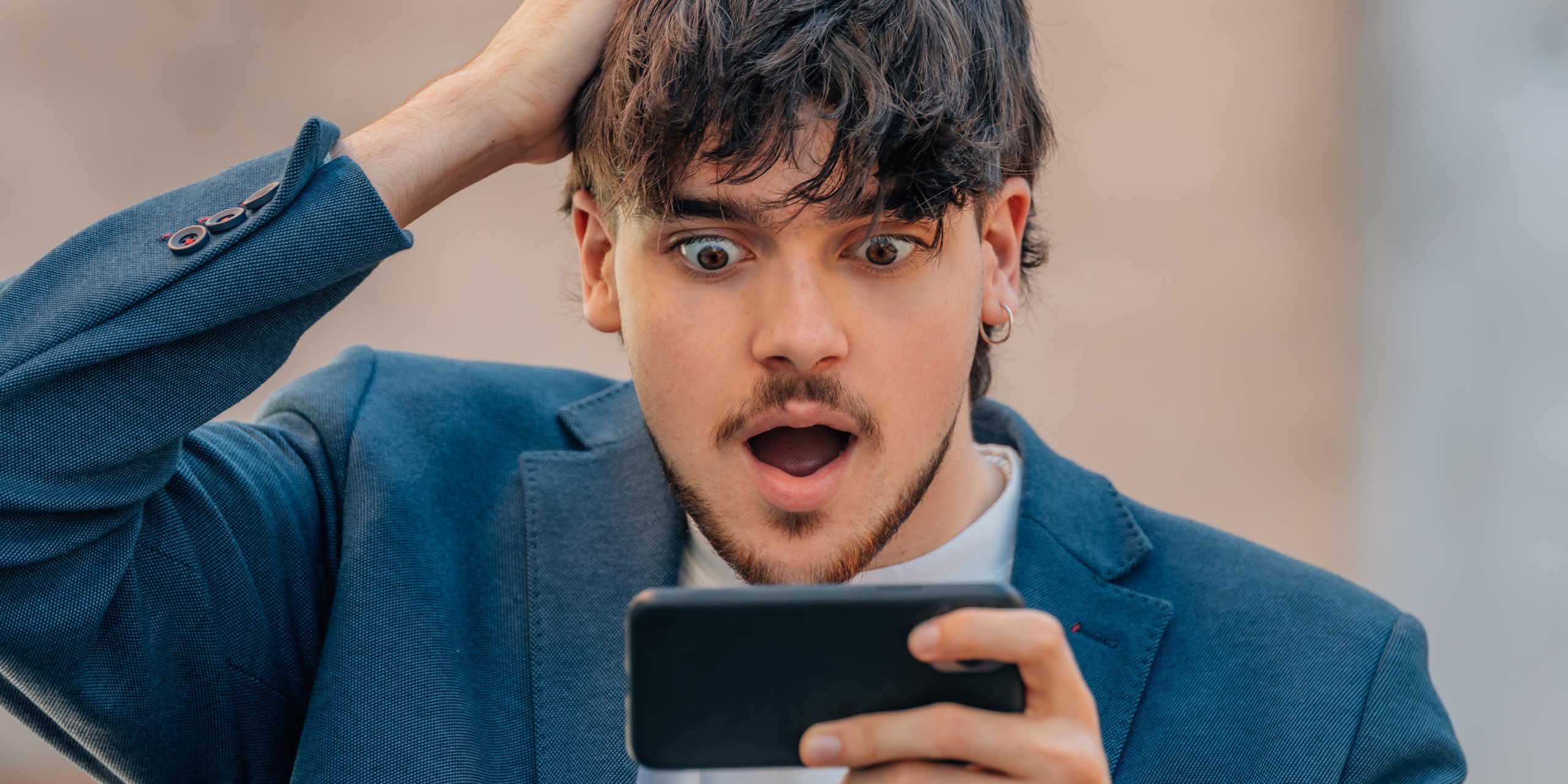 A young man places his right hand on his head while looking excitedly at his cellphone, which he is holding in his left.