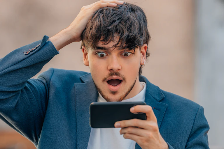 A young man places his right hand on his head while looking excitedly at his cellphone, which he is holding in his left.