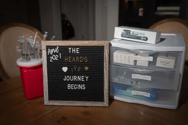A small blackboard is surrounded by medical syringes and a little plastic shelf. The board says 'The Heard's IVF journey begins.'