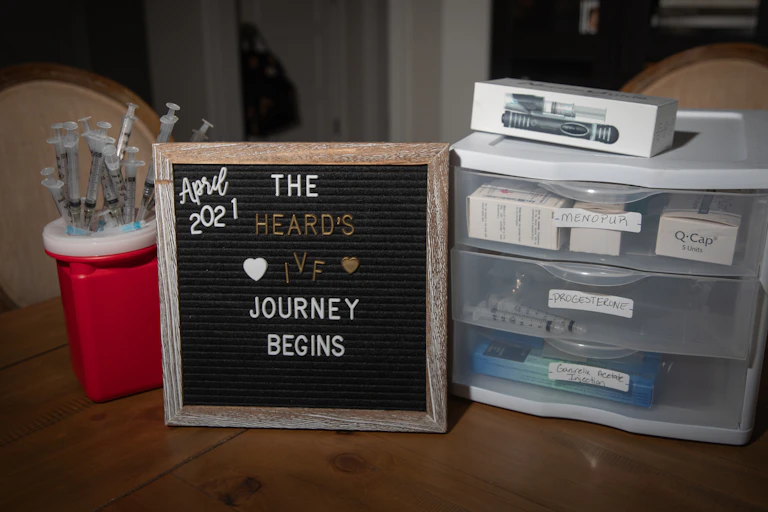 A small blackboard is surrounded by medical syringes and a little plastic shelf. The board says 'The Heard's IVF journey begins.'