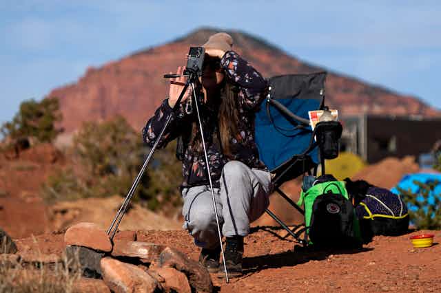 A person squatting next to a tripod holding a phone, which is aimed towards the sky. 