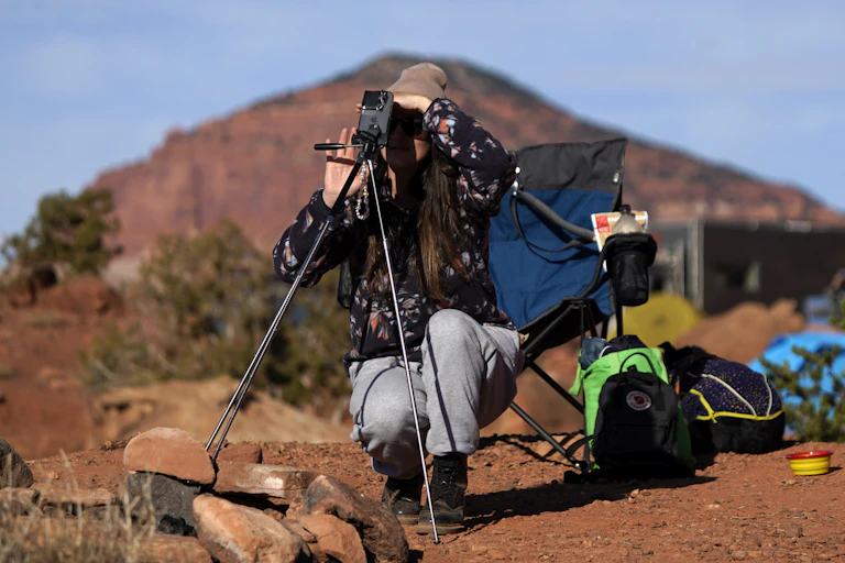 A person squatting next to a tripod holding a phone, which is aimed towards the sky.