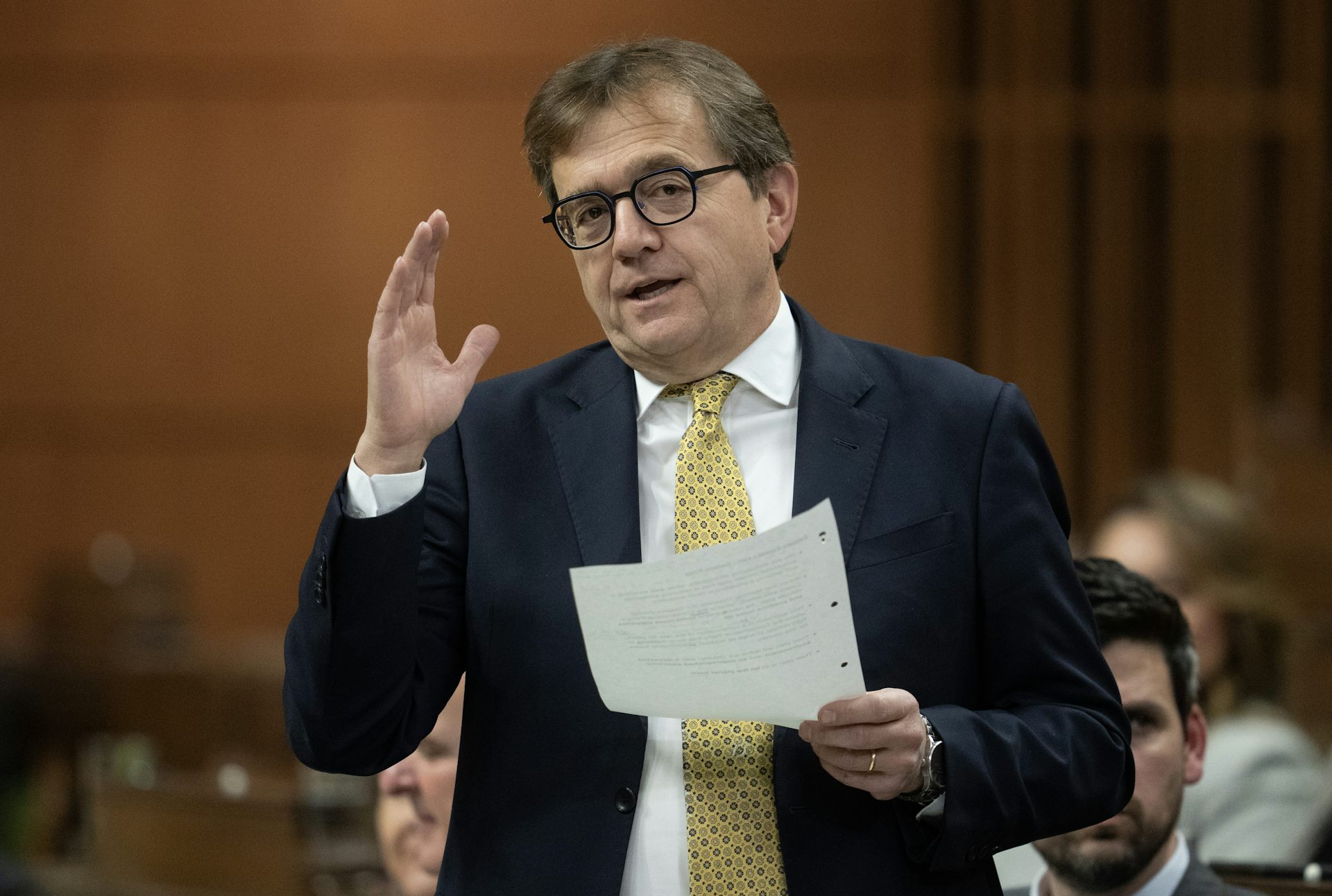 A middle-aged man in a suit and glasses gestures while speaking to a room of people